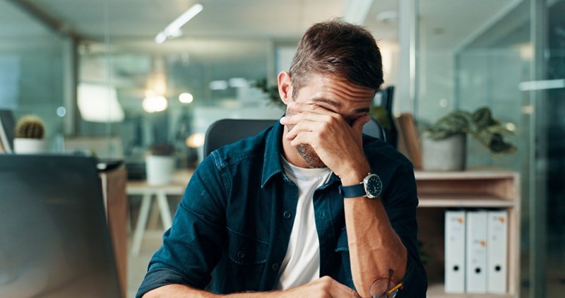 Man covering his face at an office desk with a laptop, glasses, and folders in the background.