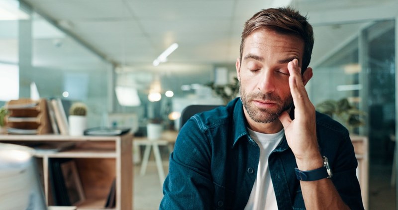 Man sitting at an office desk with his eyes closed and one hand pressed to his face.