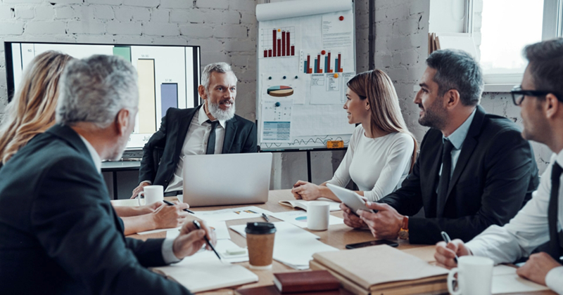 A manager sits at the head of a meeting table with a laptop, talking to his colleagues