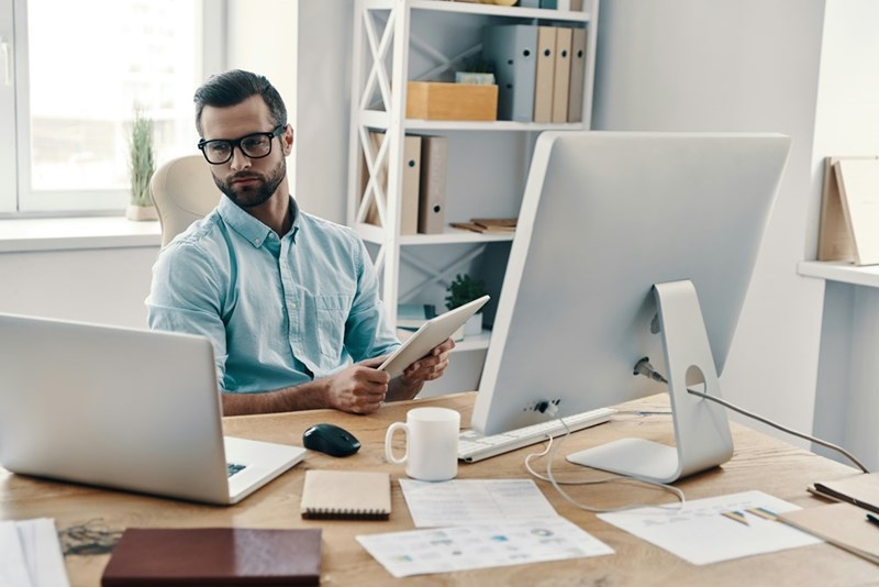 An office worker holds a tablet while seated between a laptop and a desktop computer.