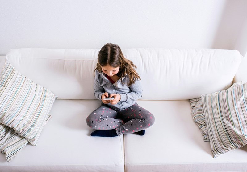 Actress depicting a girl sitting on a sofa and playing with her smartphone.