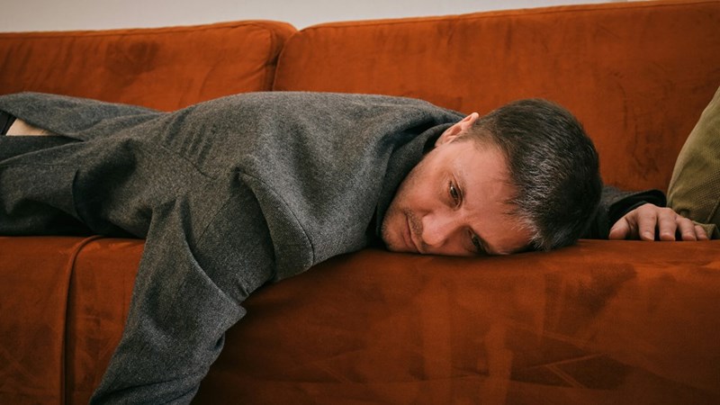 Man lying face down on an orange couch under a gray blanket, looking tired.