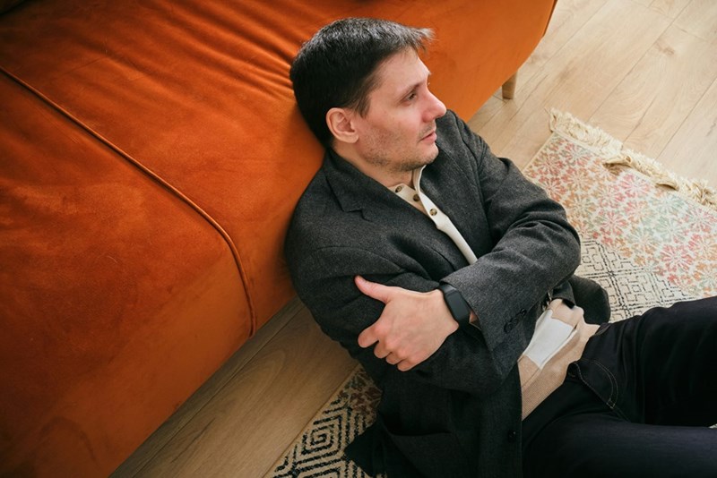 Man sitting on the floor beside an orange couch with his arms crossed, wearing a gray blazer.