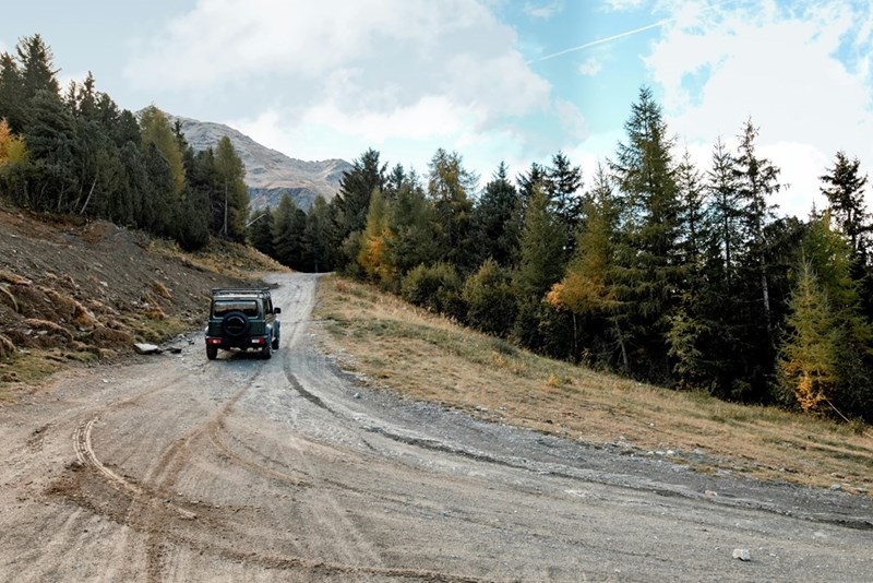 SUV driving on a rough mountain road bordered by forest and hills under a cloudy sky.