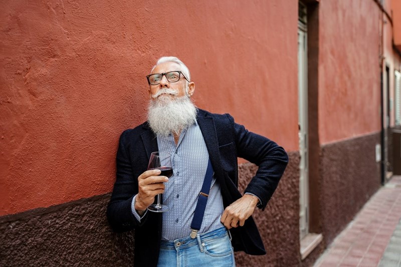An older man with a long white beard drinks a glass of wine while wearing a button-up shirt, suspenders, and blue jeans (model).
