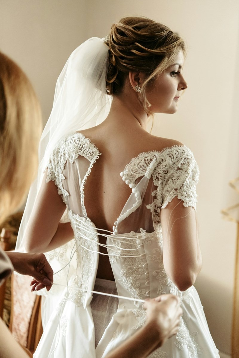 A woman gets help lacing up her wedding dress with lace and a veil, while holding a bouquet of flowers (stock).