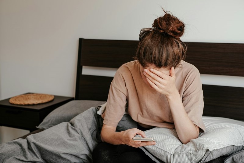 Woman wearing comfy clothes and a bun, sitting in her bed looking down at her phone.