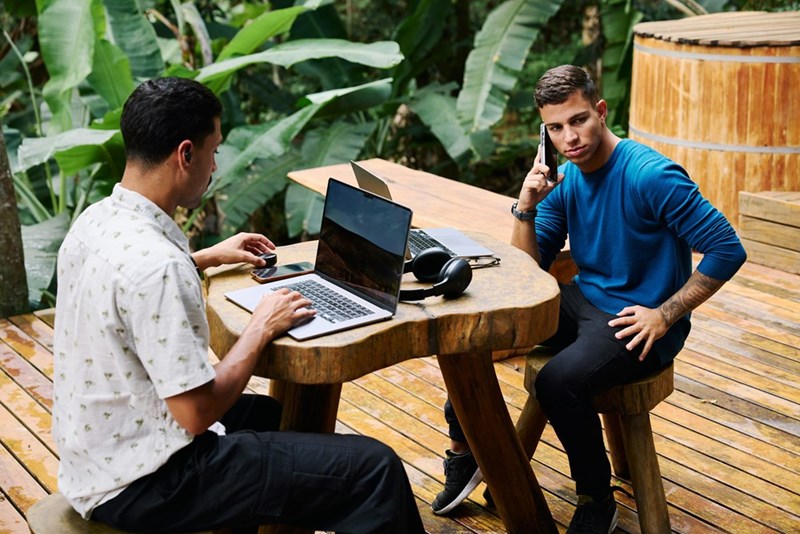 Two men sitting outside in a table with their computers and phones.