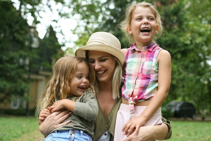 Woman wearing a hat and smiling while holding two kids.