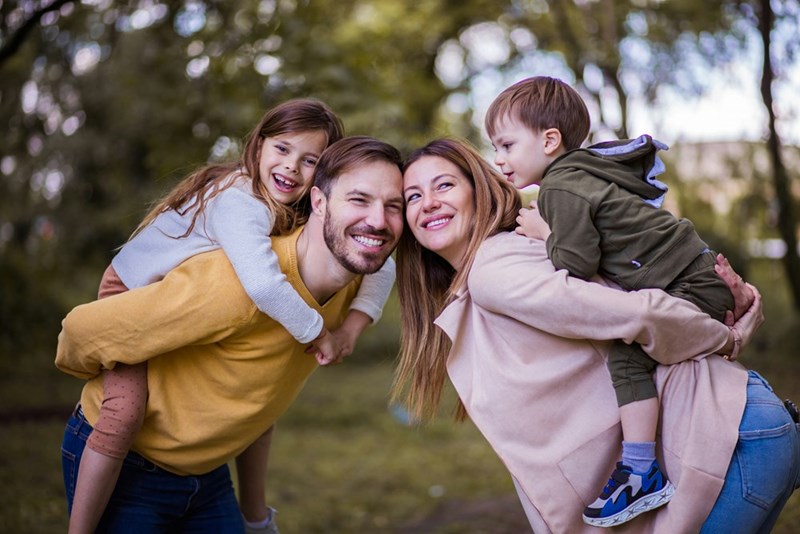 Parents holding their kid on their back smiling outside on a park.