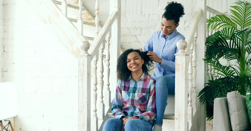 Two teenage girls sit on a staircase, with one braiding the other's hair