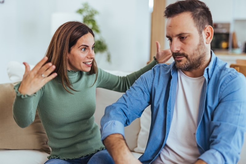 Woman gesturing while speaking to a man sitting beside her on a couch in a bright living room.