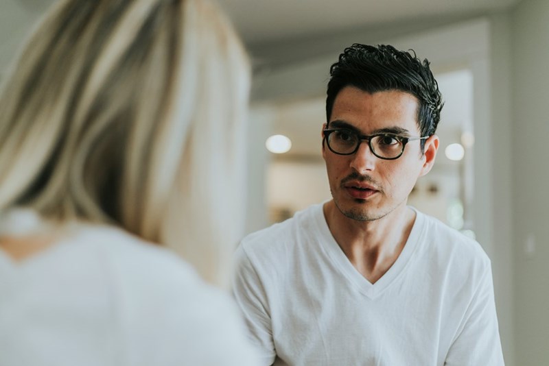 Man in glasses speaking to a woman in a bright home interior, wearing a white V-neck shirt.