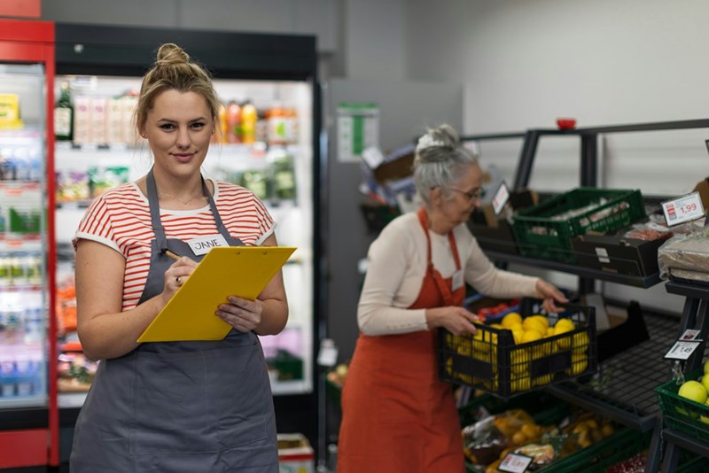 Retail manager oversees store inventory while team member stocks fresh produce shelves.