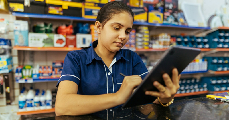 A female retail employee writes on a clipboard while leaning behind a counter