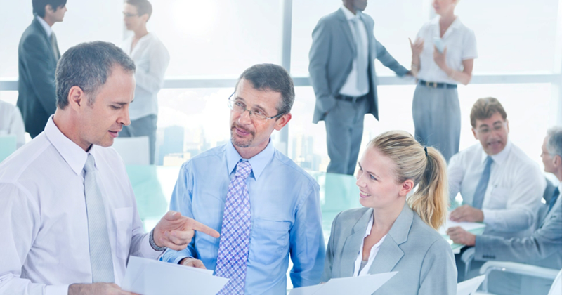 Three employees have a discussion over documents as coworkers walk around in the background