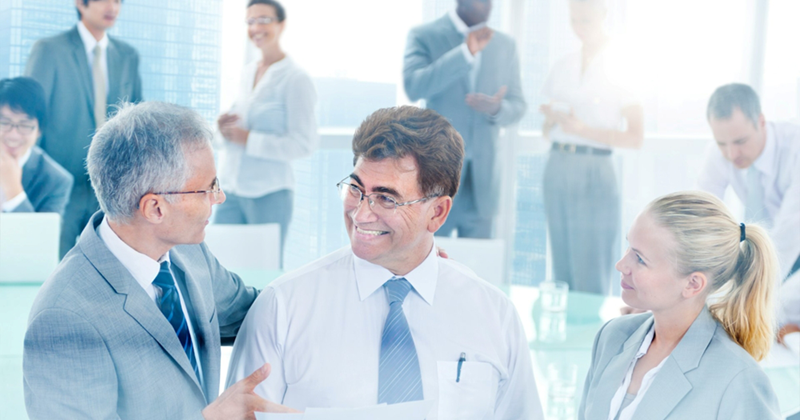 Three employees have a discussion as coworkers walk around in the background