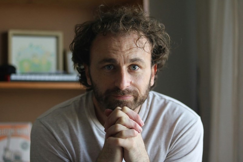 Man with curly hair sitting indoors with clasped hands, looking at the camera in soft light.