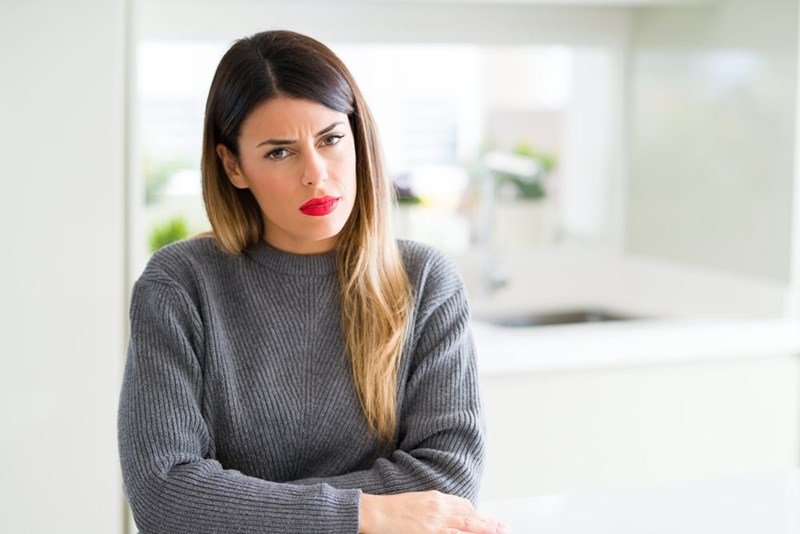 Woman in a gray sweater sits with crossed arms and a skeptical expression in a bright kitchen.