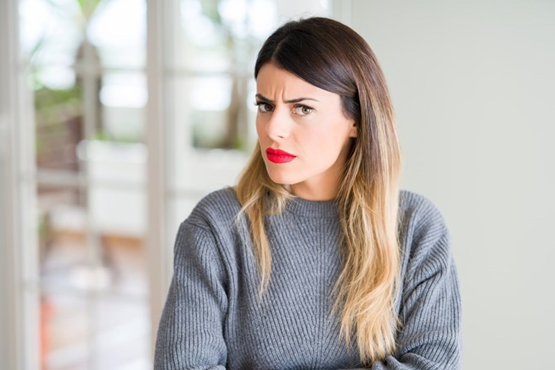 Woman in a gray sweater looks sideways with a skeptical expression in a bright room.