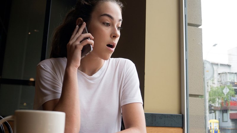 Woman in a white shirt talks on a phone by a window with a surprised expression and coffee mug nearby.