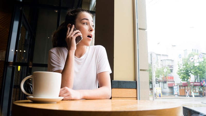 Woman in a white shirt talks on a phone at a cafe table by a window, with a coffee mug nearby.