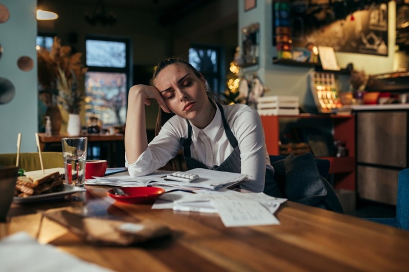 Tired woman wearing an apron rests her head on her hand at a cafe table covered with papers and a calculator.