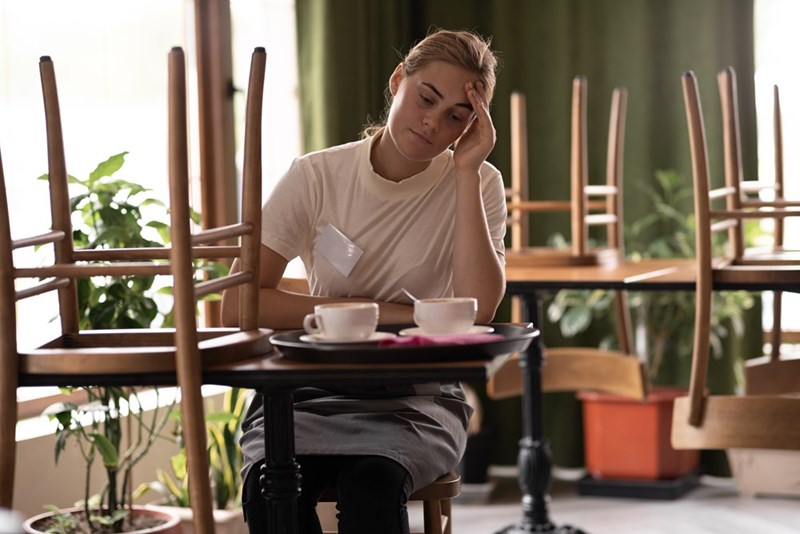 Woman sits with her hand on her forehead among upside-down cafe chairs, beside a tray with cups.