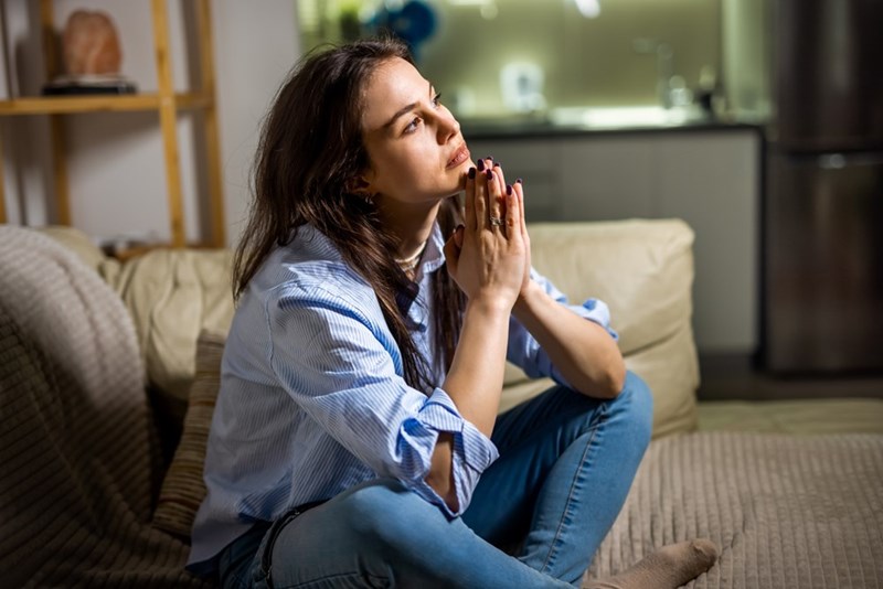 Thoughtful woman sits on a sofa with clasped hands, looking upward in a modern living room.