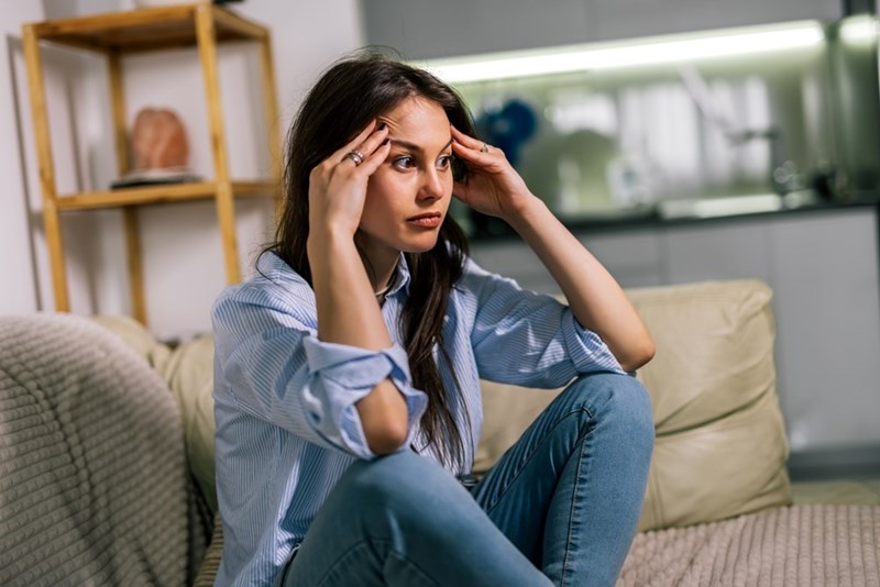 Worried woman sits on a sofa with her hands on her temples in a modern living room.