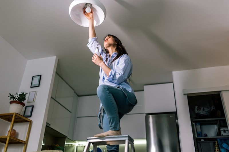 Woman standing on a step stool changes a ceiling light bulb in a modern kitchen.