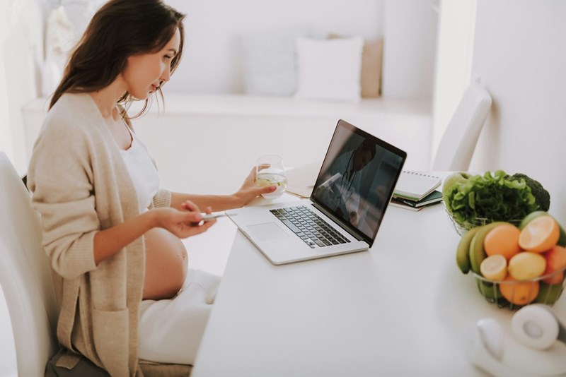 Pregnant woman sitting down in a chair looking at her computer lying on the table.