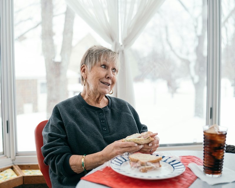 Old woman eating a sandwich in a kitchen table.