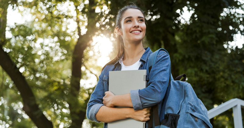 A young female college student smiles as she stands outside, holding a notebook to her chest