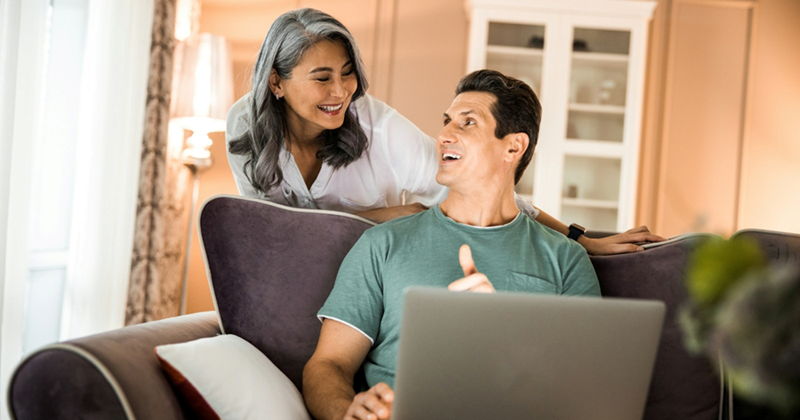 A man sits on a couch and uses a laptop, looking up at a woman smiling behind him