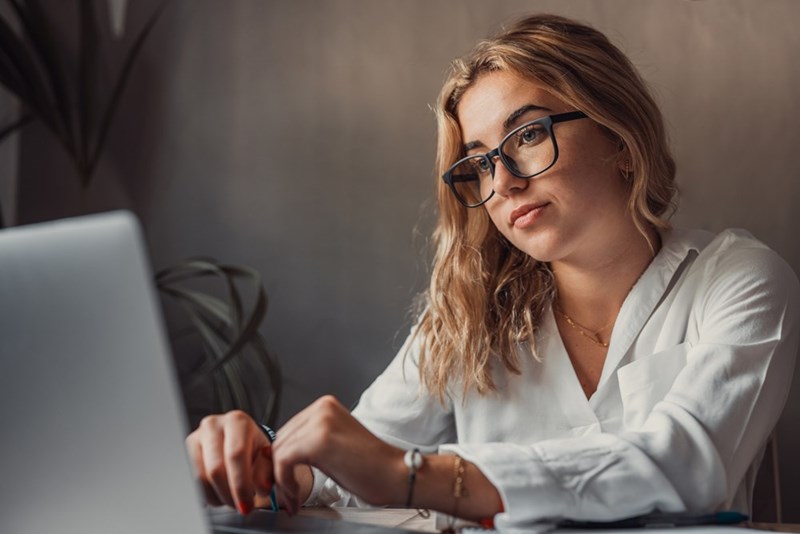 Woman wearing glasses works at a laptop in a white blouse with a serious expression.
