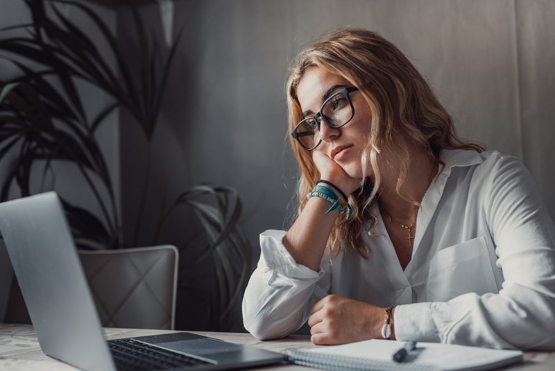 Woman wearing glasses rests her cheek at a desk beside a laptop and notebook.