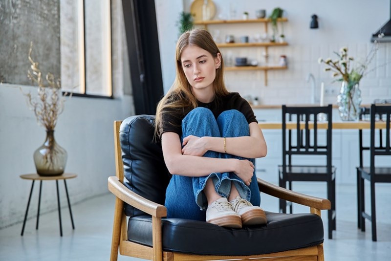 Young woman sitting curled up in an armchair in a bright room with a kitchen behind her.