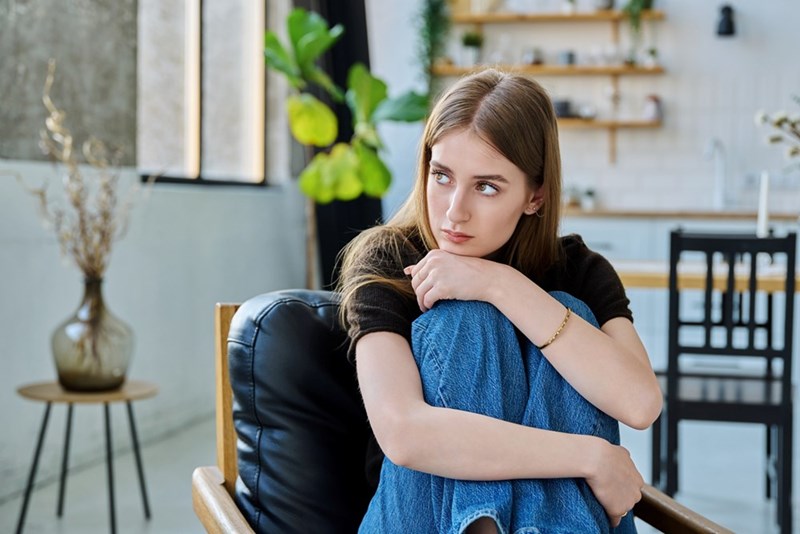Young woman sitting curled up in an armchair, hugging her knees in a bright room with a kitchen behind her.