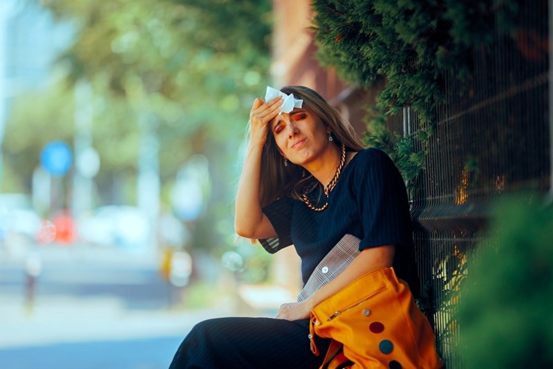 Woman sitting outdoors with a tissue on her forehead, wearing a black dress beside an orange bag.