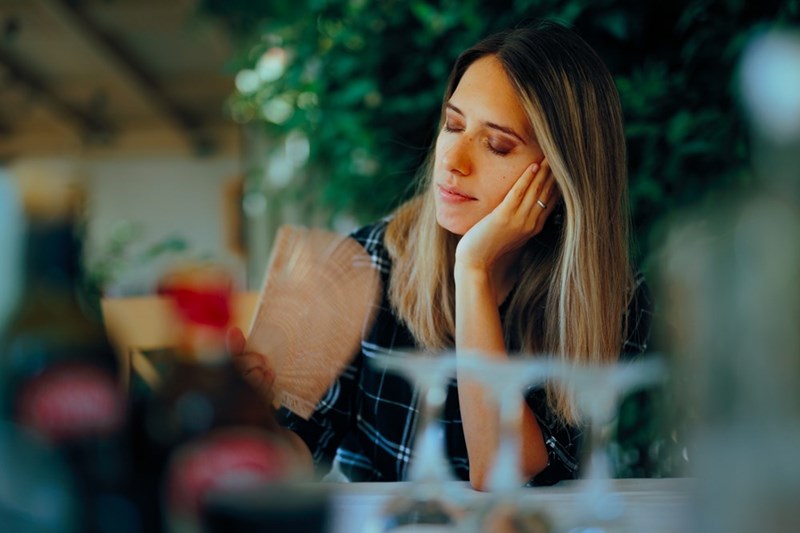 Woman resting her cheek in her hand while holding a paper fan at a table with blurred glasses in front.