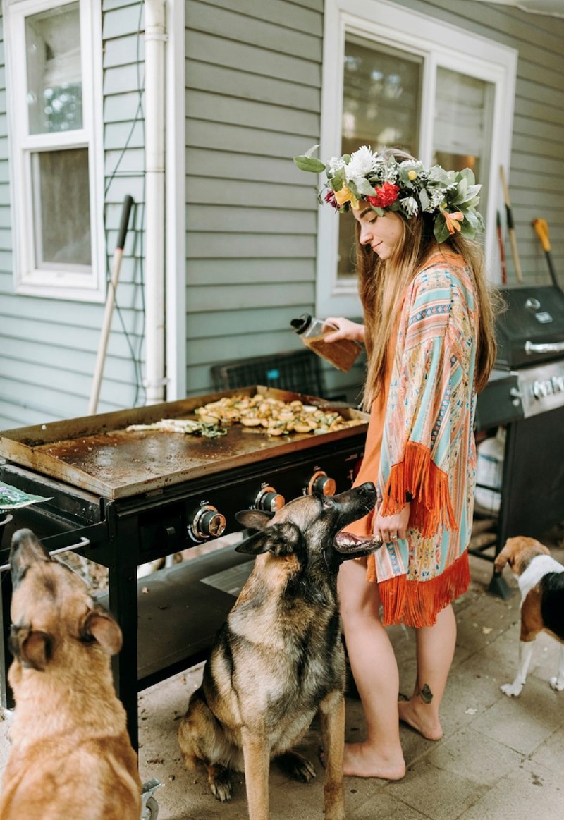 A woman in an elegant flower crown cooks at the barbecue at a 4th of July party while her two loyal German shepherds wait for her and some food.