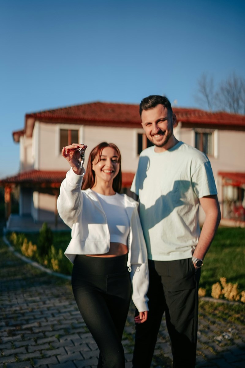 New homeowners standing in front of their house and holding up their keys after completing the purchase of the property.