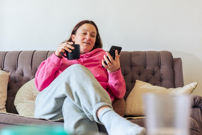 Woman on a couch with a phone and mug, looking amused but unimpressed, like someone’s complaint is not changing her mind