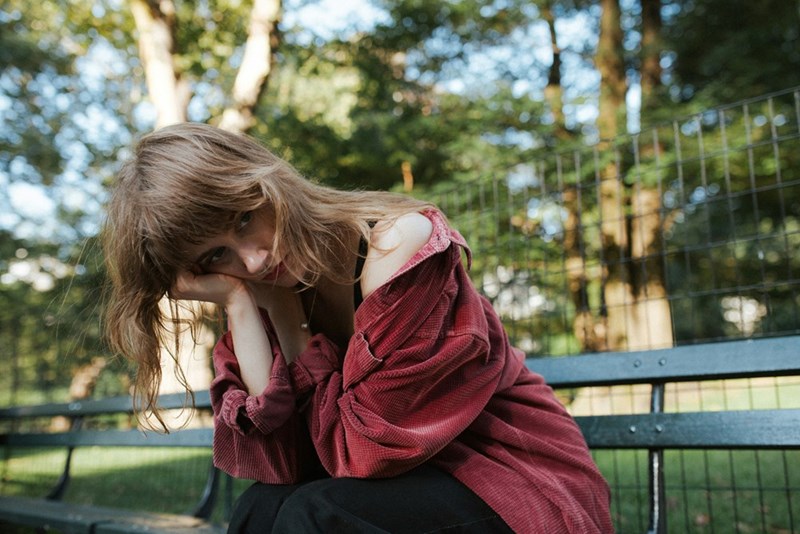 Young woman sitting on a park bench in a red overshirt, resting her cheek on her hand.