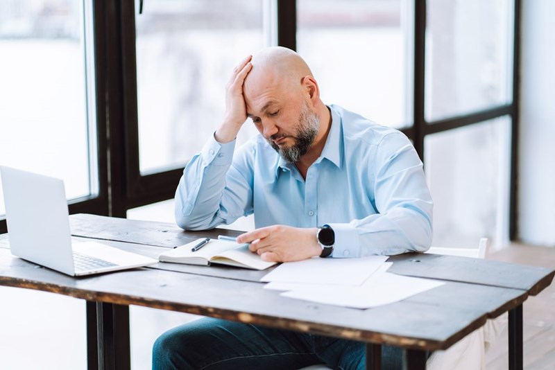 Man sitting at a table with laptop, notebook, and papers, holding his head near large windows.