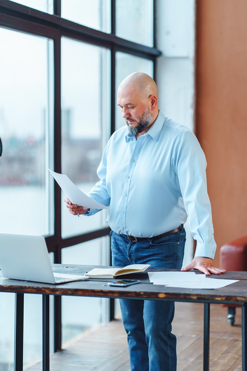 Man standing by a table with a laptop and papers, reading a document near large windows.