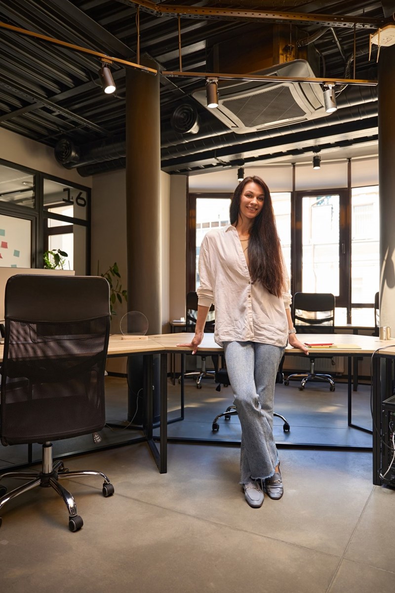 Woman standing in a modern office beside desks and chairs, wearing a white shirt and jeans.