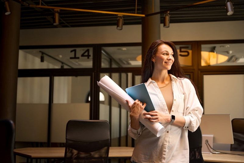 Woman smiling in a modern office while holding rolled papers and a blue folder near desks and glass doors.