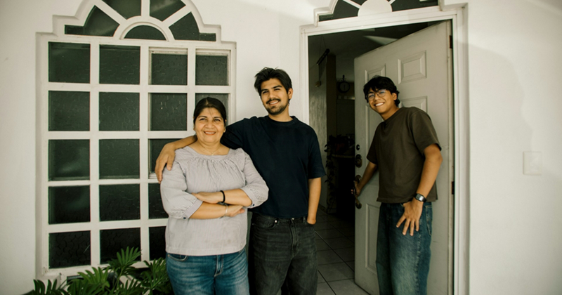 A man smiles as he opens the front door to a house, as another man puts his arm around the shoulder of a woman beside him